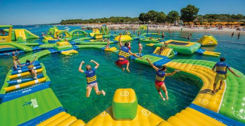 A vibrant water park with inflatable structures, people jumping into the water, and a sunny beach in the background.
