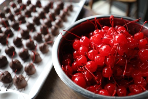 A bowl of bright red cherries with stems next to a tray of chocolate-covered treats.
