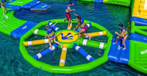 A colorful inflatable water park with people jumping and playing on floating structures in clear blue water.