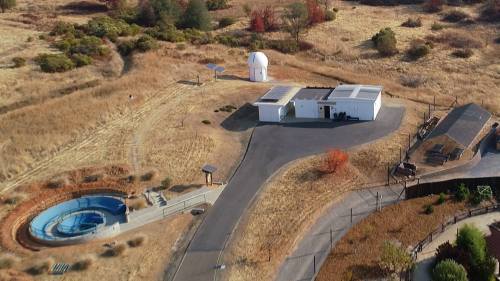 Aerial view of a facility with a dome structure, surrounded by dry grass and trees, featuring a pool and solar panels.