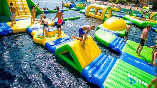 Children and teens play on colorful inflatable water slides and obstacles in a sunny outdoor pool area.