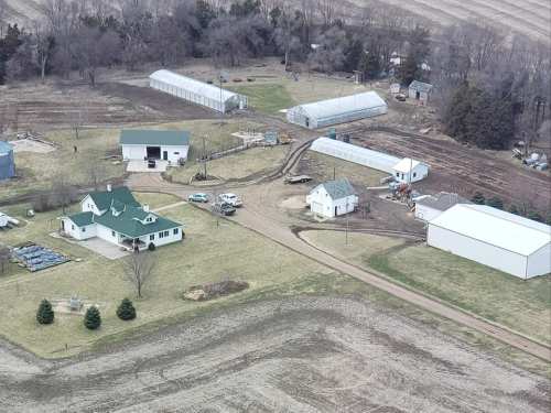 Aerial view of a rural farm with greenhouses, a house, and several outbuildings surrounded by fields.