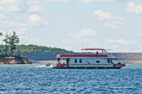 A houseboat floats on a calm lake, with a jet ski in the background and trees lining the shore.