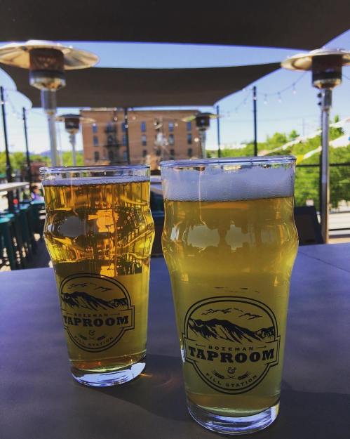 Two pints of beer sit on a table at an outdoor taproom, with a sunny backdrop and patio heaters in view.