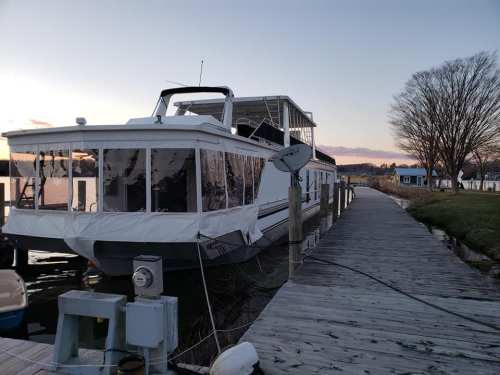 A houseboat docked at a wooden pier during sunset, with trees and buildings in the background.