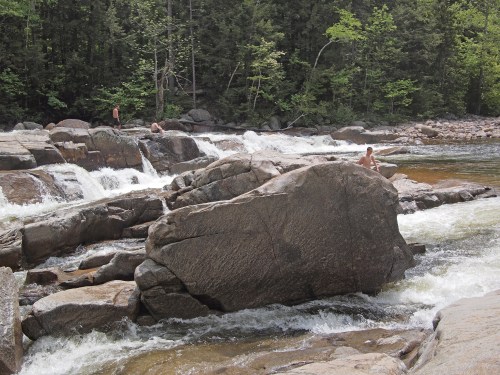 People enjoying a rocky river landscape with flowing water and lush green trees in the background.
