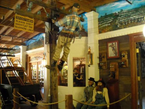 A mannequin climbs a wooden pole in a museum, with vintage decor and other mannequins seated nearby.