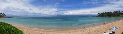A panoramic view of a serene beach with clear blue water, sandy shore, and a few people enjoying the sun.