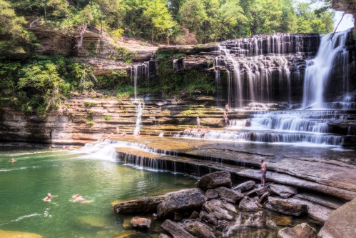 A scenic waterfall cascades into a serene pool, with people swimming and enjoying the natural beauty around them.