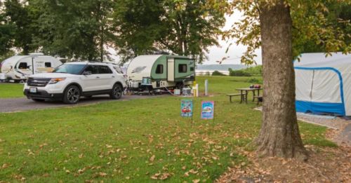 A campsite with a green trailer, an SUV, and a blue tent, surrounded by trees and grass, near a lake.
