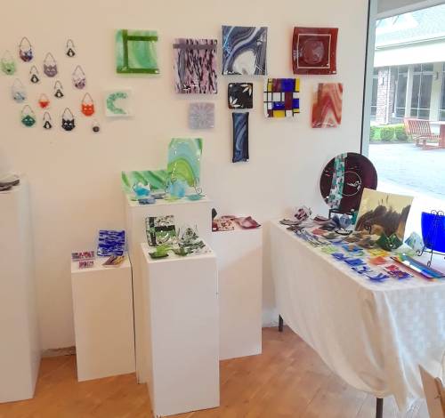 Art gallery display featuring colorful glass art pieces on white pedestals and walls, showcasing various designs and styles.
