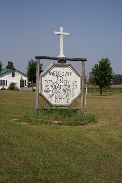 Sign welcoming visitors to Tyewhoppety, KY, with a population of 33, featuring a cross and a message to bless America.