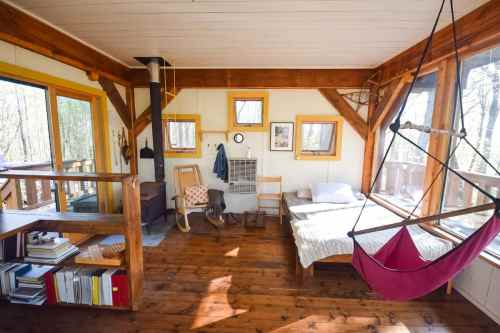 Cozy cabin interior with wooden beams, a hammock, bed, and a small wood stove, surrounded by large windows.