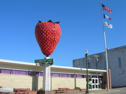 A large strawberry statue stands in front of a building with flags flying, representing Strawberry Point. Clear blue sky.