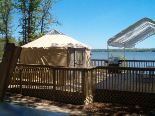 A lakeside view featuring a tent and a covered patio with a grill, surrounded by trees and clear blue skies.