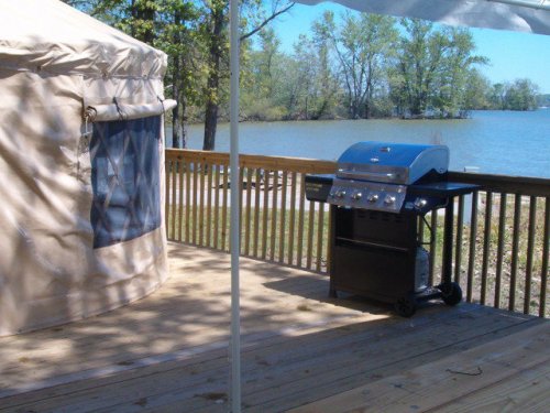 A wooden deck by a lake featuring a blue grill and a tan yurt under a white canopy. Trees line the water's edge.