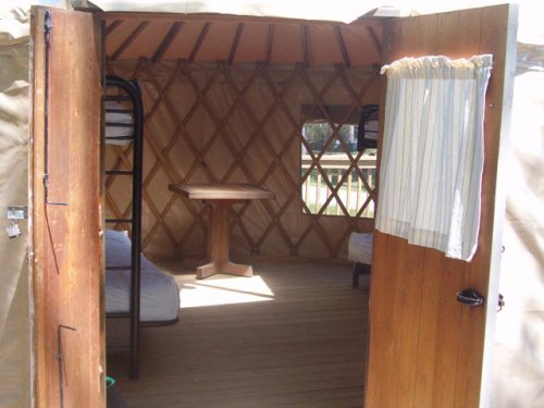 Interior of a yurt with wooden doors, a central table, and two beds against the walls. Natural light enters through the door.