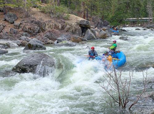 A group of people in a blue raft navigating a turbulent river surrounded by rocky terrain and trees.