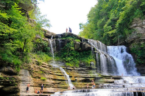 A scenic waterfall surrounded by lush greenery, with people swimming and enjoying the water at its base.