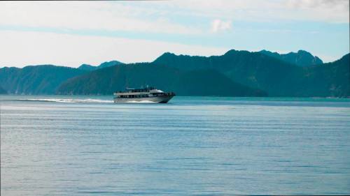A ferry boat glides across calm waters, with mountains in the background under a clear sky.