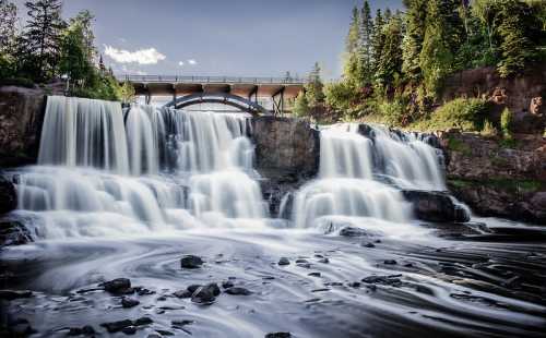 A scenic waterfall cascades over rocks, with a bridge in the background and lush greenery surrounding the area.