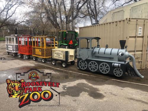 A colorful miniature train at Roosevelt Park Zoo in Minot, North Dakota, parked near a green tractor.