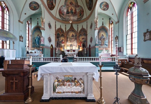 Interior of a church featuring an altar with a white cloth, stained glass windows, and ornate religious artwork.