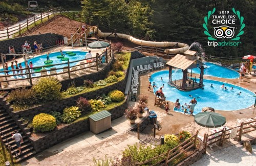 Aerial view of a water park with pools, slides, and families enjoying the sun. Lush greenery surrounds the area.