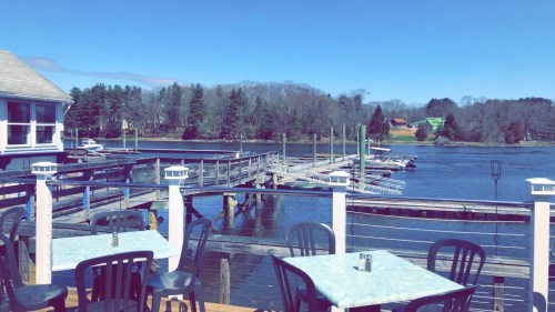 A scenic view of a calm lake with docks, boats, and trees under a clear blue sky from a waterfront restaurant.
