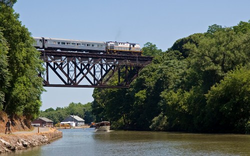 A train crosses a tall bridge over a river, with trees and a small building visible along the bank.