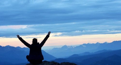 A person sits on a rock with arms raised, overlooking a mountain landscape at sunset.