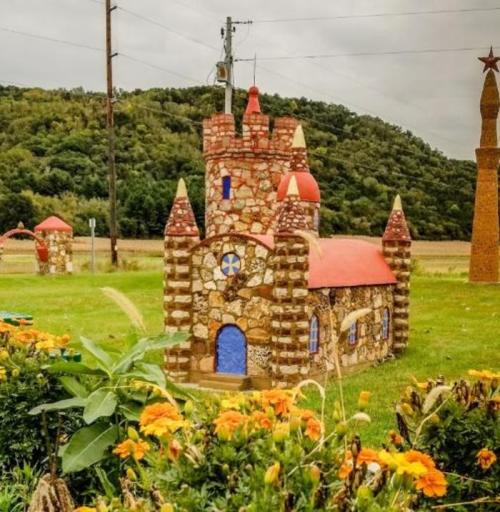 Colorful stone castle with red roofs surrounded by flowers and greenery, set against a hilly landscape.