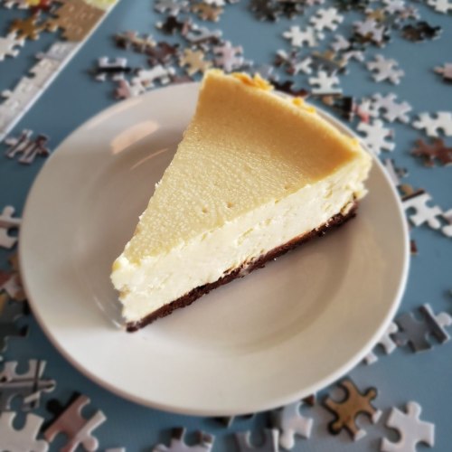 A slice of cheesecake on a white plate, surrounded by scattered puzzle pieces on a blue table.