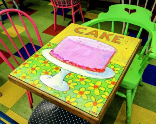 A colorful table with a painted pink cake and the word "CAKE" surrounded by flowers, set in a vibrant café.