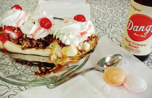 A banana split topped with whipped cream, cherries, and nuts, alongside a bottle of red cream soda and a spoon.