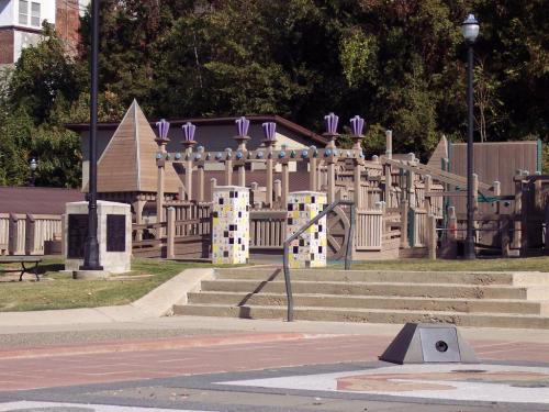 A wooden playground with colorful decorations, surrounded by trees and a pathway leading to it.