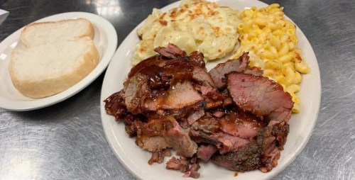 A plate of sliced smoked meat with barbecue sauce, macaroni and cheese, cheesy potatoes, and two slices of bread.
