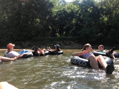 A group of people relaxing on inner tubes in a river, surrounded by trees and sunlight.
