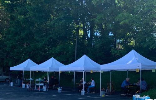 A row of white tents in a parking lot, with people seated at tables under the shade of trees.