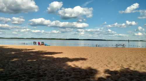 A sandy beach with calm water, dotted with clouds in the sky and a few people enjoying the scenery.