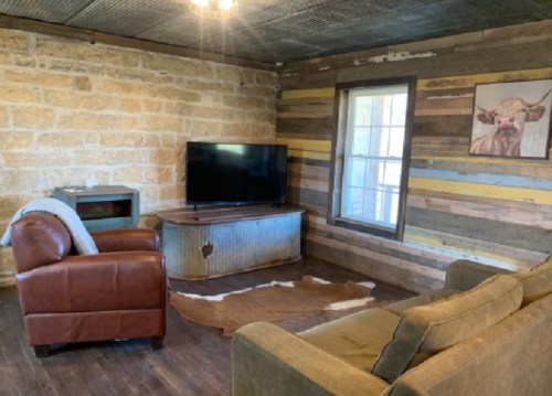 Cozy living room with stone walls, a TV on a metal stand, a brown leather chair, a green sofa, and a cowhide rug.