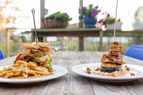 Two gourmet dishes on white plates: a towering sandwich with fries and a stacked seafood dish, set on a rustic wooden table.
