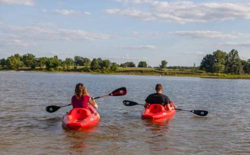 Two people in red kayaks paddle on a calm lake, surrounded by greenery and a blue sky with scattered clouds.