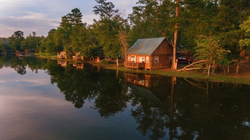 A serene lakeside scene featuring wooden cabins surrounded by trees, reflecting in calm water at sunset.