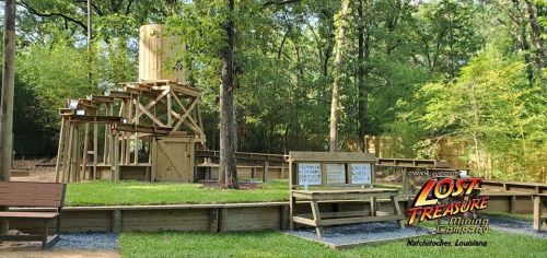 A wooden mining structure surrounded by trees, with benches and informational signs in a park setting.