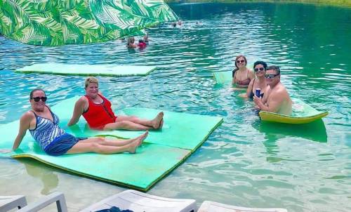 A group of five people relax on floating mats in a pool, enjoying a sunny day surrounded by water.