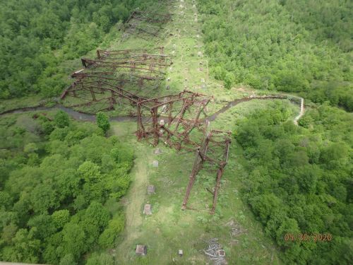 Aerial view of a rusted, collapsed structure surrounded by lush green trees and a winding creek.