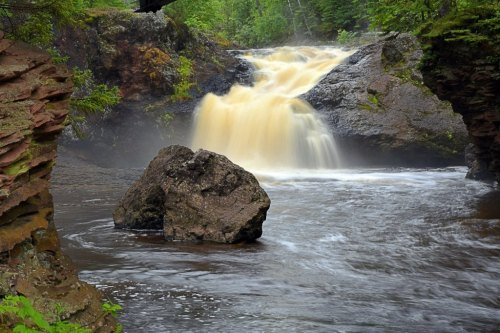 A serene waterfall cascades over rocks into a calm river, surrounded by lush greenery and mist.