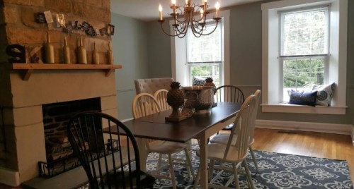 A cozy dining room with a wooden table, chairs, a chandelier, and a stone fireplace, featuring natural light from windows.