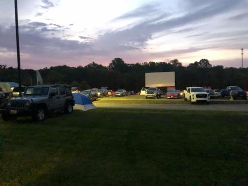A drive-in movie scene at dusk, with cars parked and a large screen illuminated against a colorful sky.